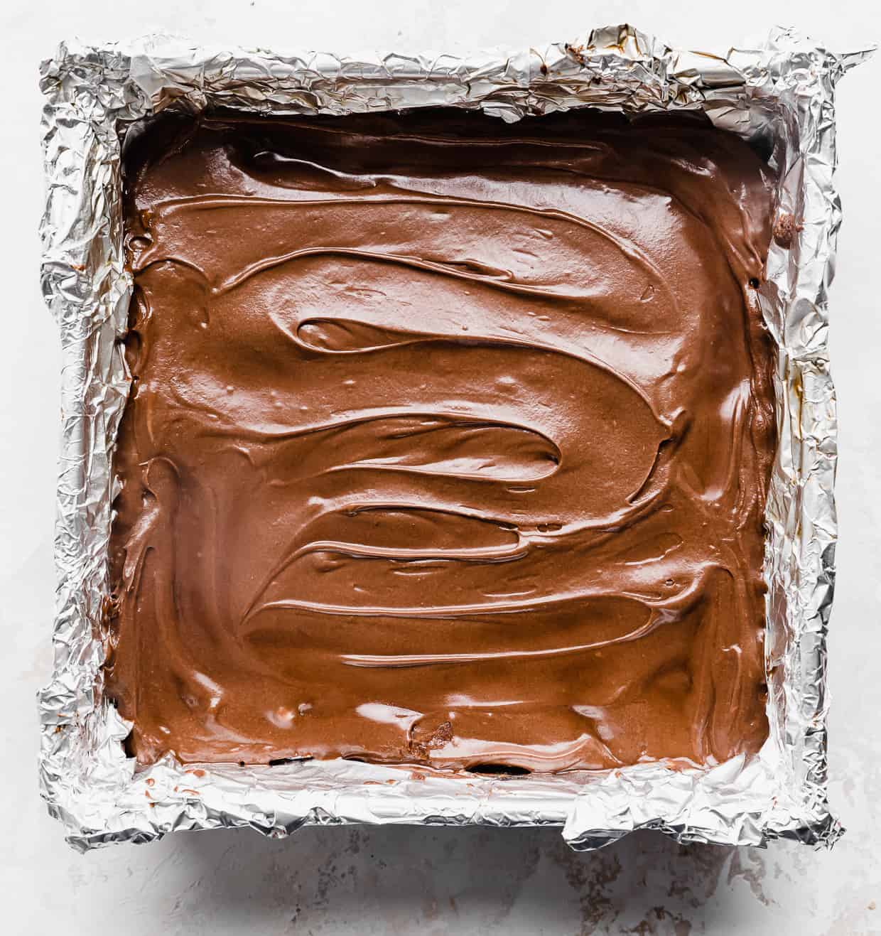 Freshly frosted lunch lady brownies in a foil lined square pan against a white background.