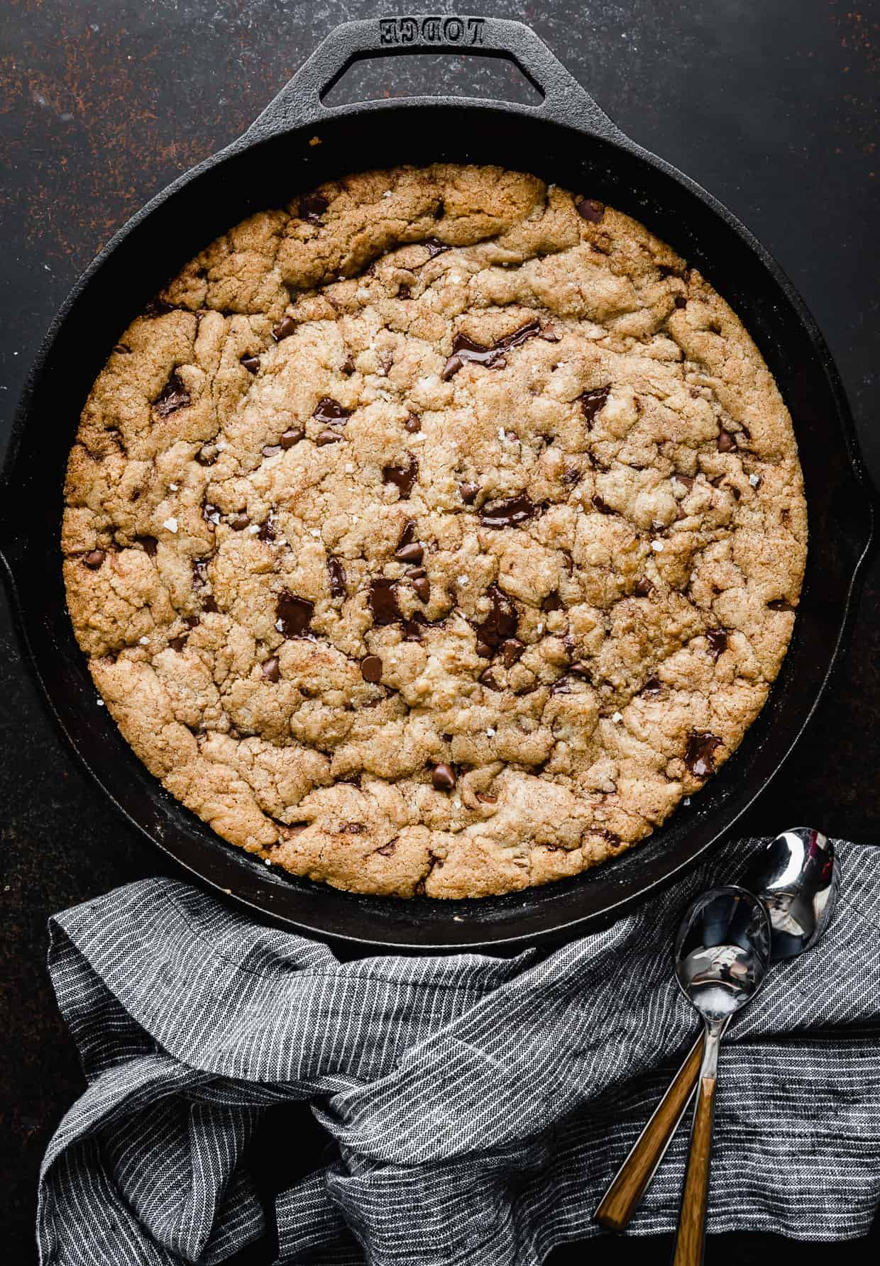 A chocolate chip cookie baked in a large cast iron skillet, against a dark brown background.