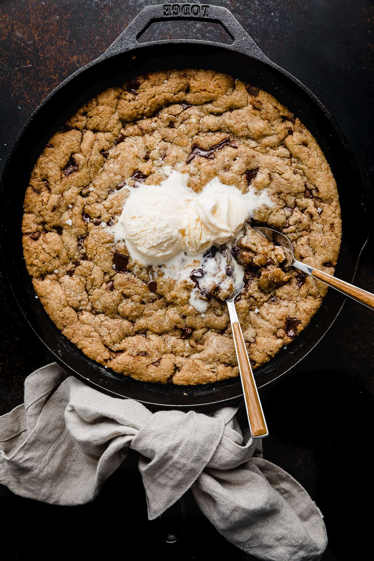 Slightly melty vanilla ice cream scooped on top of a large skillet chocolate chip cookie (pizcokie) with two spoons stuck in the center. 