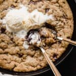 Two spoons scooping up skillet chocolate chip cookie that has been topped with vanilla ice cream.