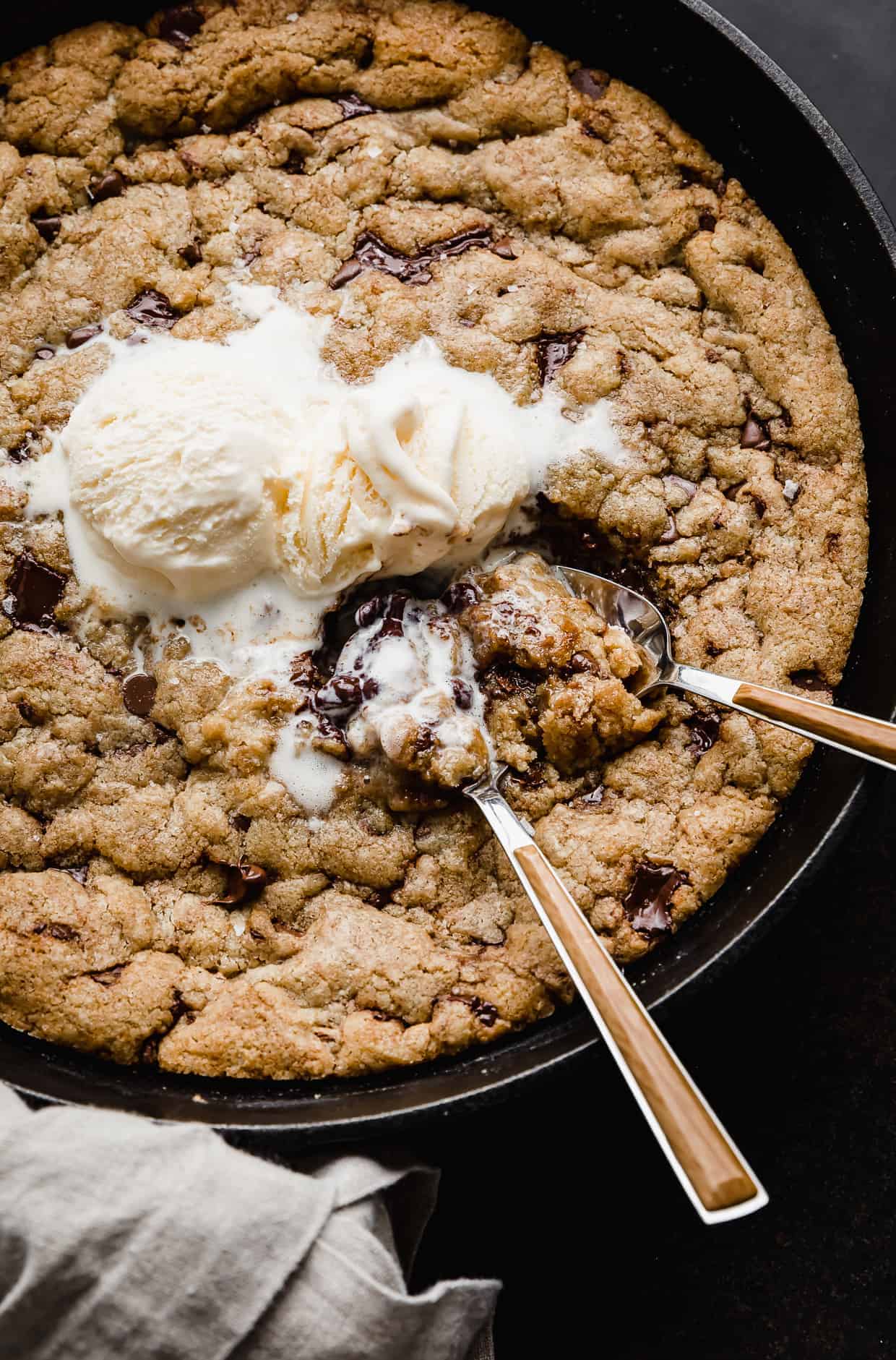 Two spoons scooping up skillet chocolate chip cookie that has been topped with vanilla ice cream. 
