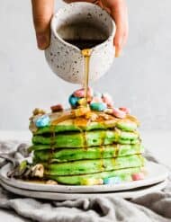 Maple syrup being poured overtop Lucky Charms topped green pancakes for St. Patrick's Day.