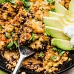 A One Pan Mexican Quinoa recipe with a serving spoon scooping into the Mexican quinoa skillet topped with avocado slices.