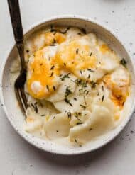 A Creamy Scalloped Potatoes Recipe in a white bowl topped with fresh thyme and cheese.