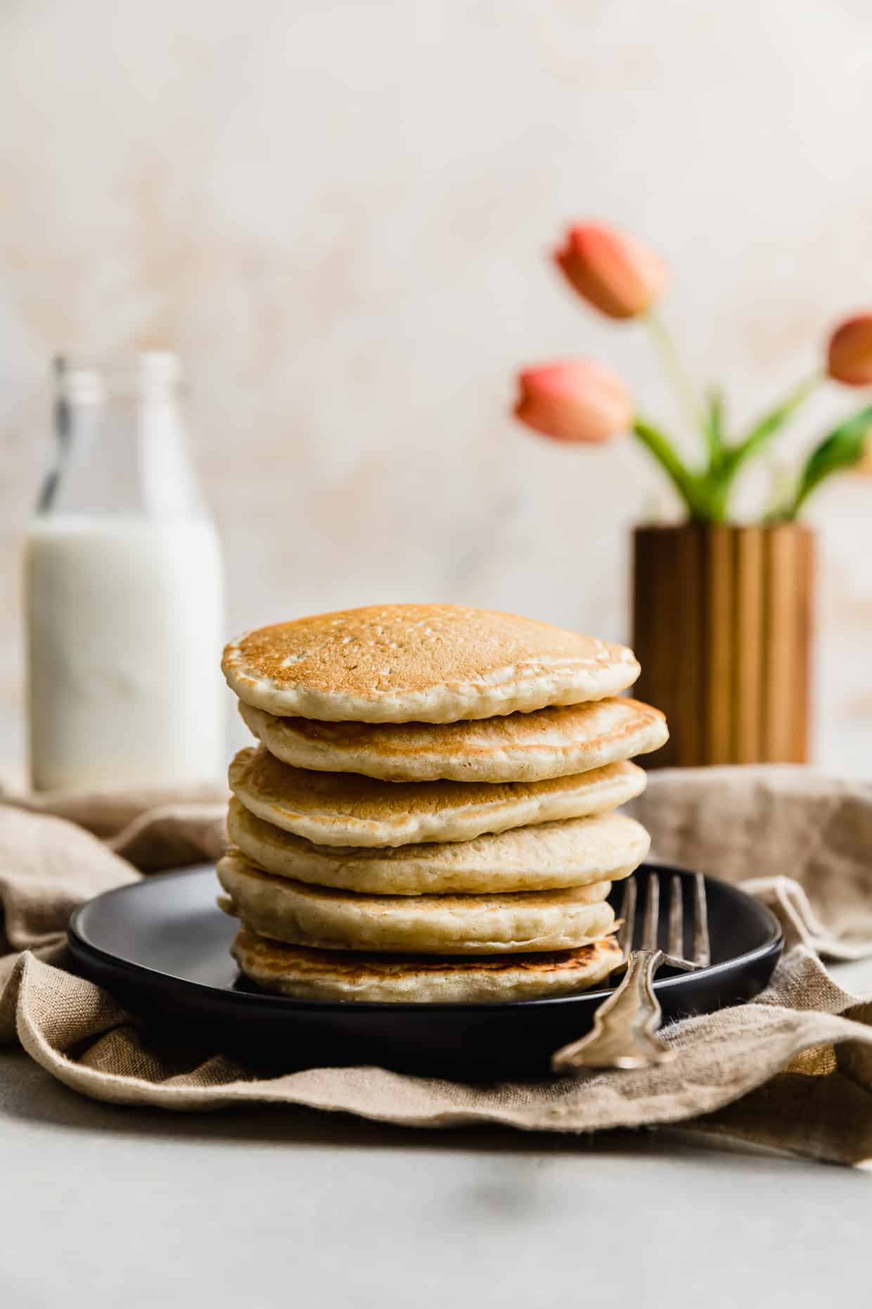 A stack of Almond Milk Pancakes on a black plate with pink tulips in the background.