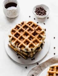 A stack of square Chocolate Chip Waffles on a white plate with syrup in the background.