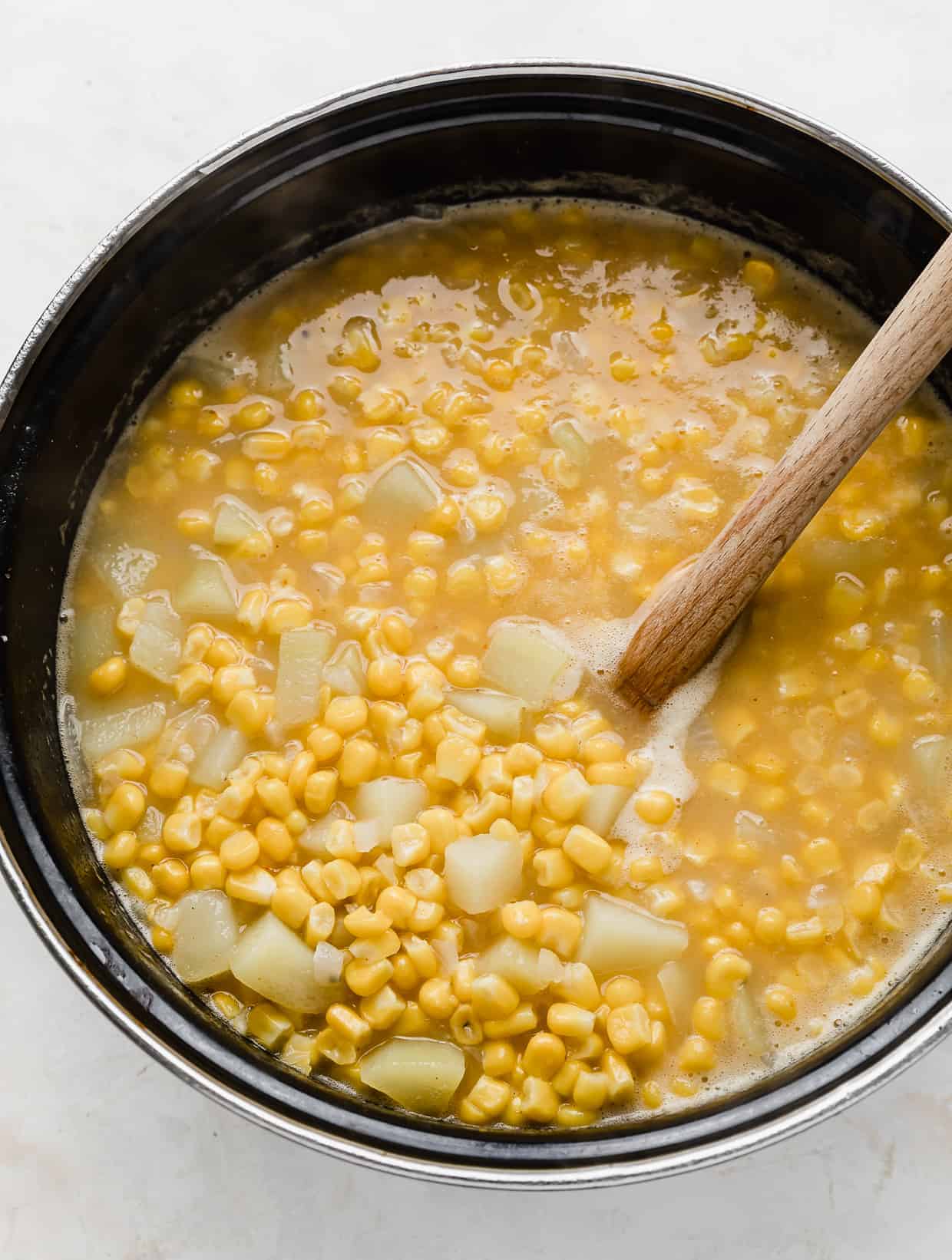 A wooden spoon stirring a large pot full of cooked corn and diced potatoes.