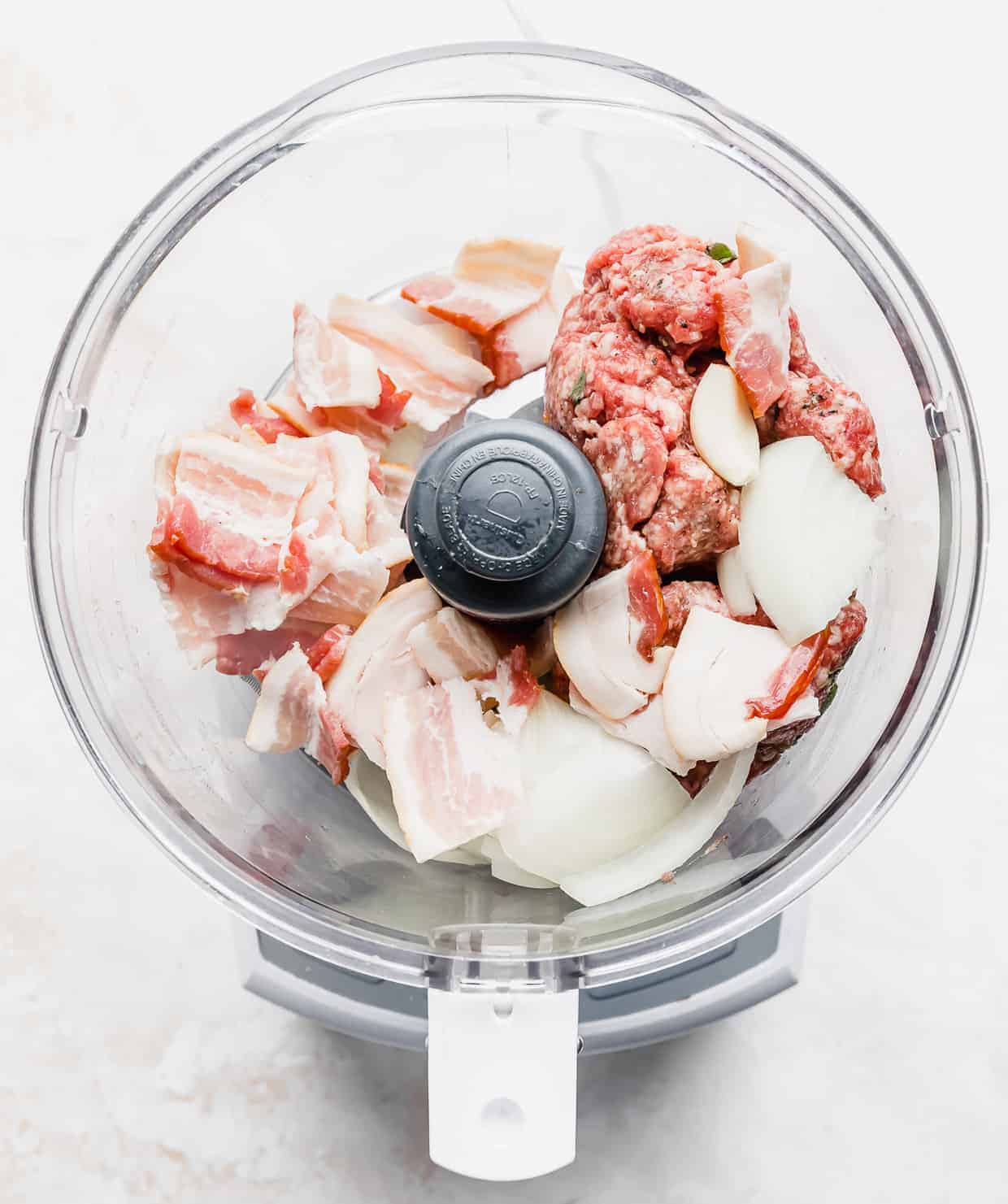 A food processor with raw bacon, onion, and ground lamb in it.