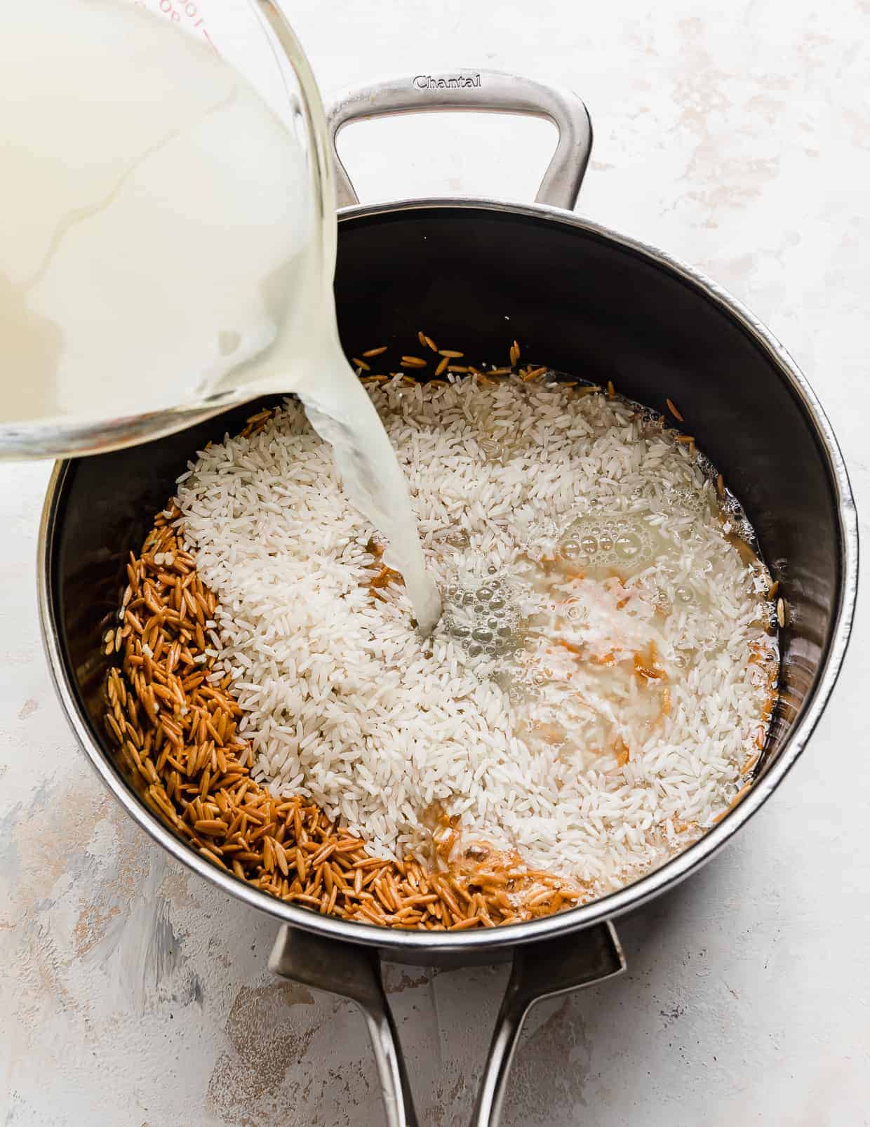 Chicken broth being poured overtop of white rice and orzo in a saucepan.