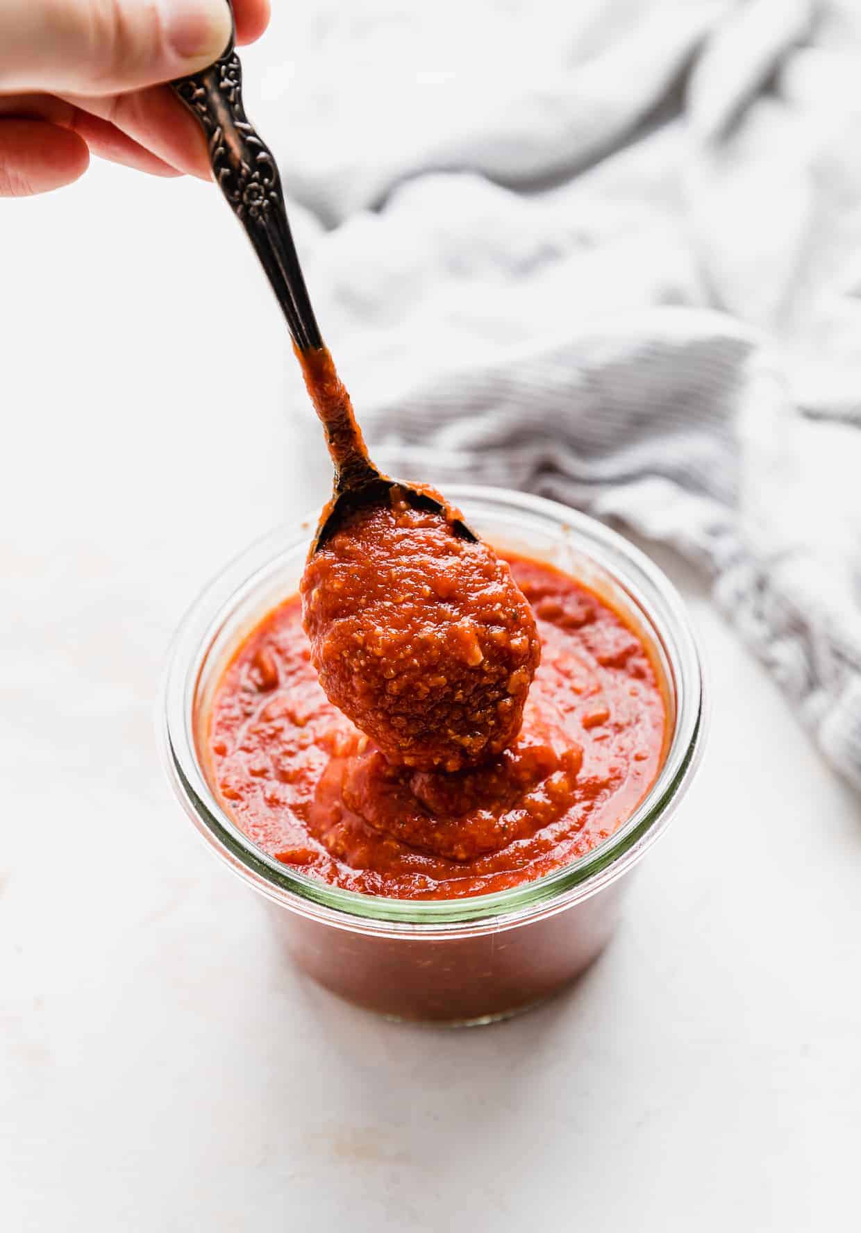 A spoon scooping up homemade pizza sauce from a glass jar. 