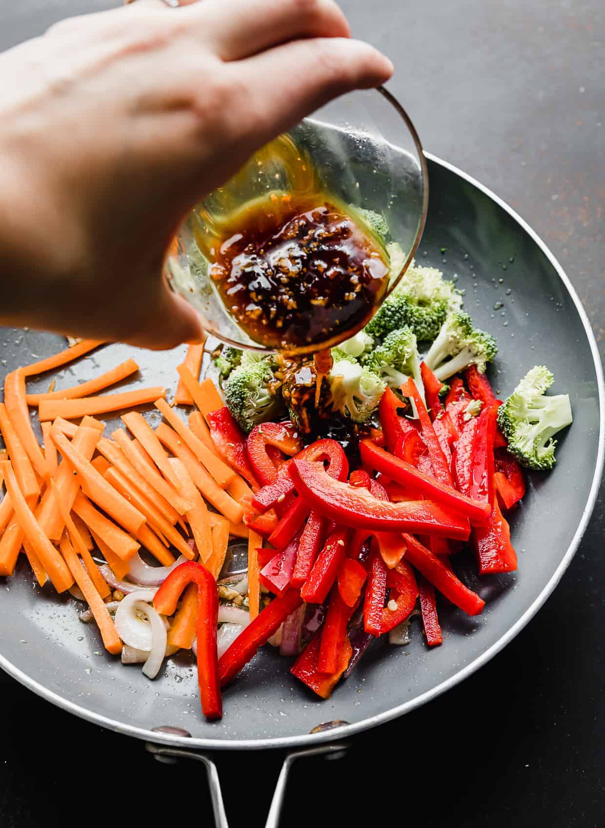A hand pouring a brown sauce overtop of veggies in a skillet.