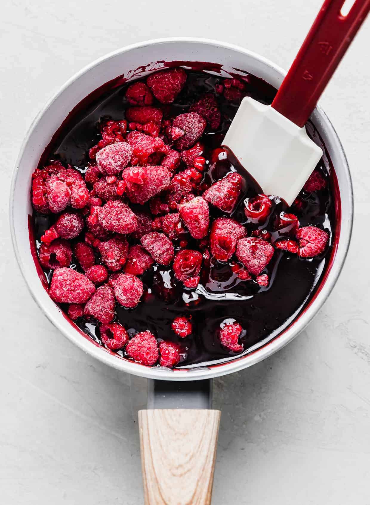Frozen raspberries being stirred into a red sauce in a pot.