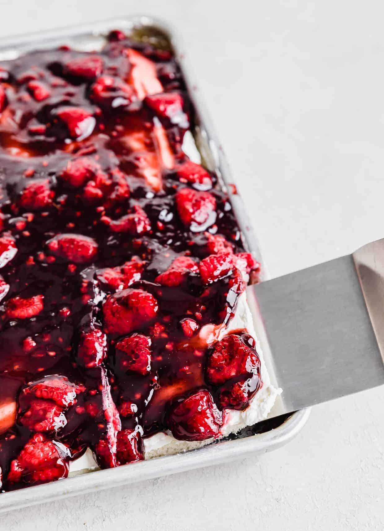 A metal spatula lifting up a slice of Raspberry Shortcake Sheet Cake from a baking sheet. 