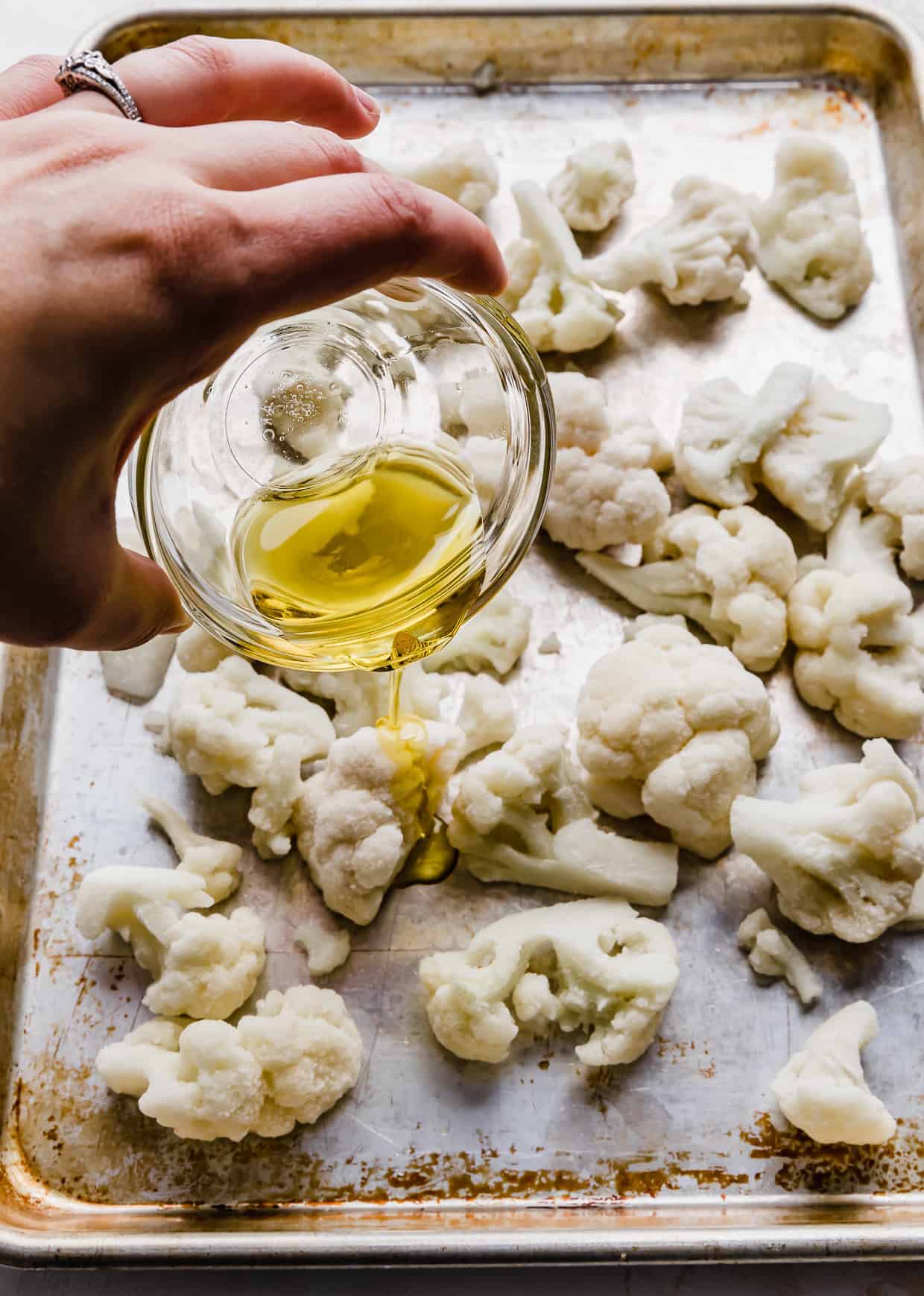 Olive oil being poured over frozen cauliflower on a baking sheet.