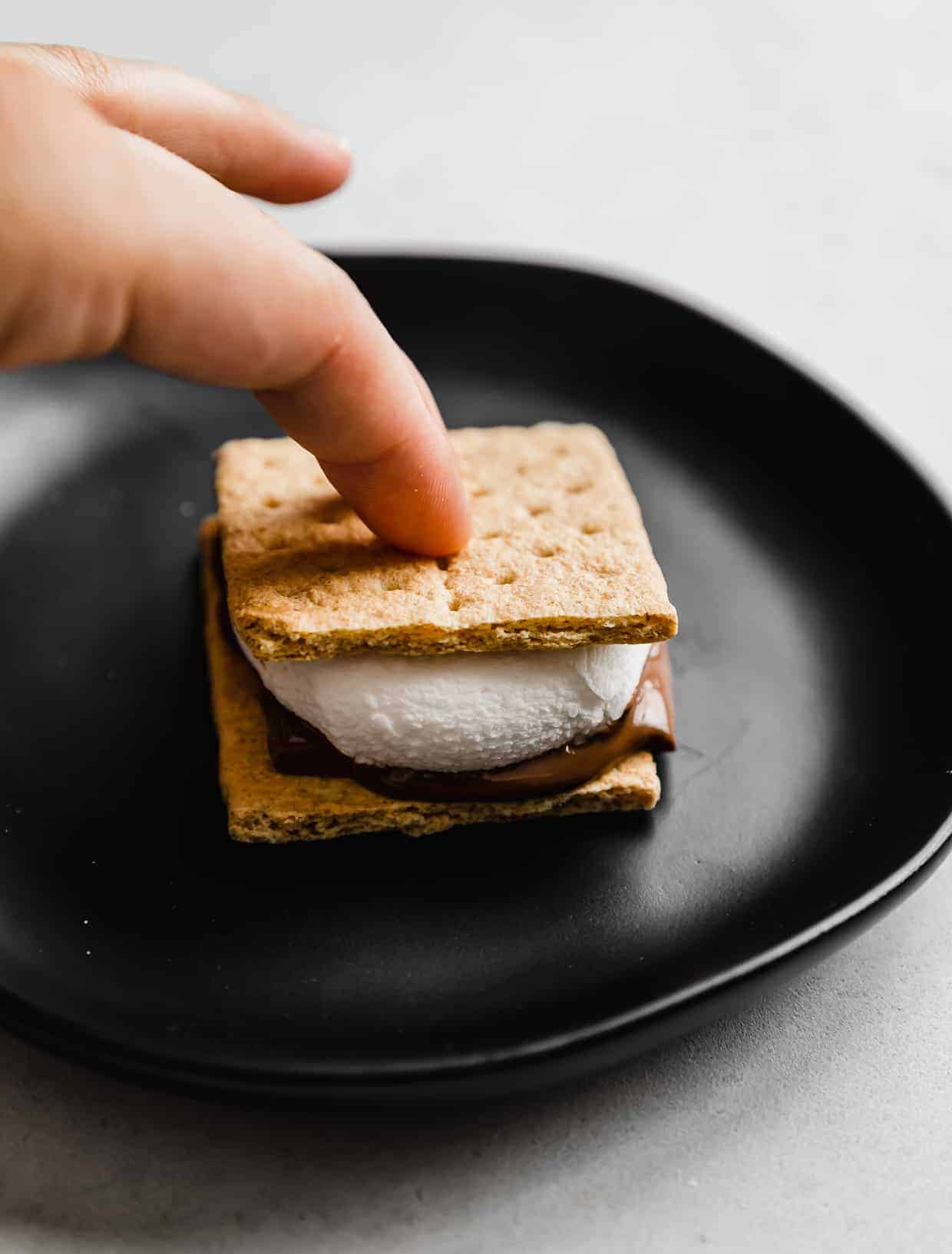 A finger pressing a graham cracker square on top of a large marshmallow to make a s'more.