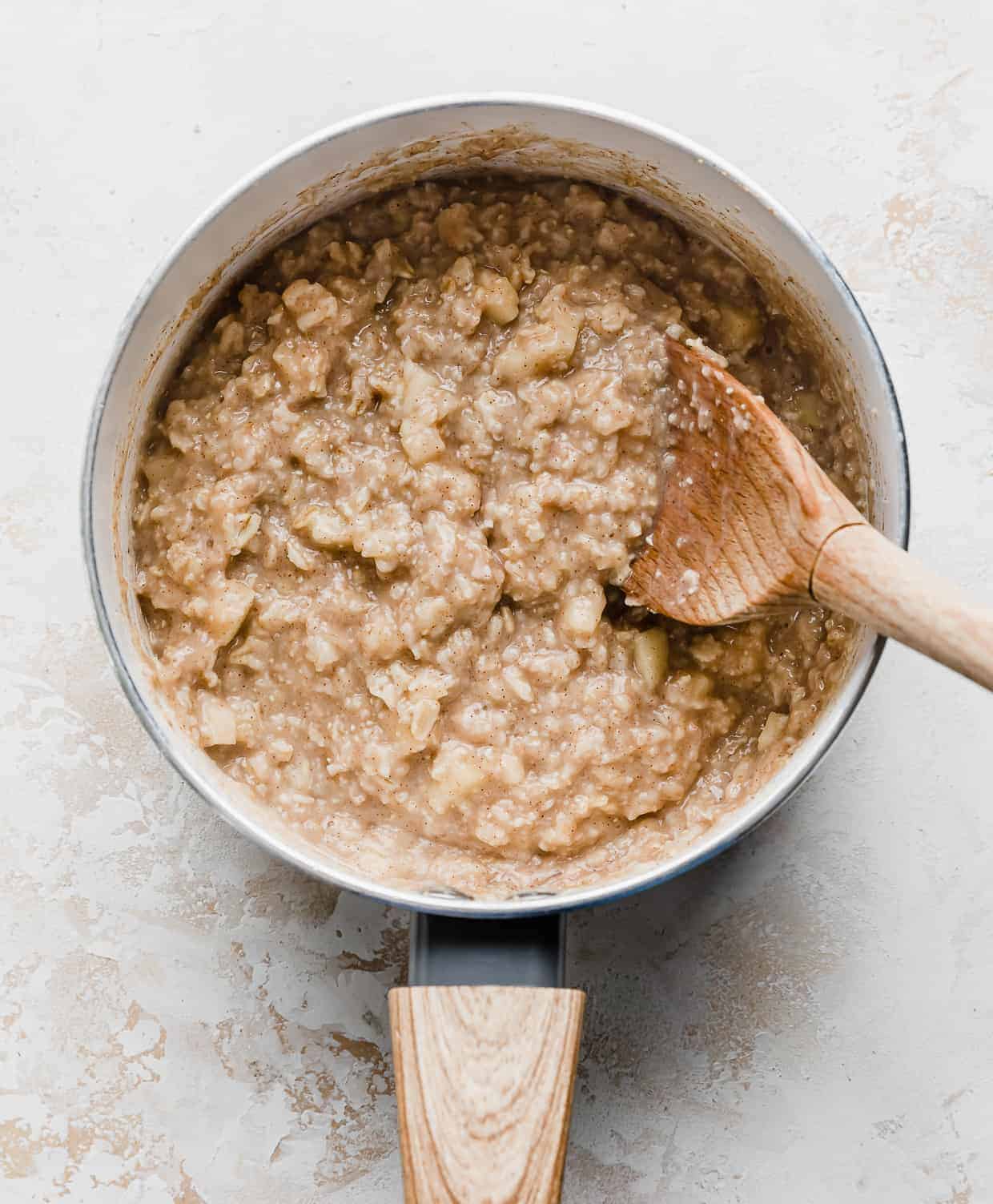 A wooden spoon inside a white pot full of cooked Apple Cinnamon Oatmeal.