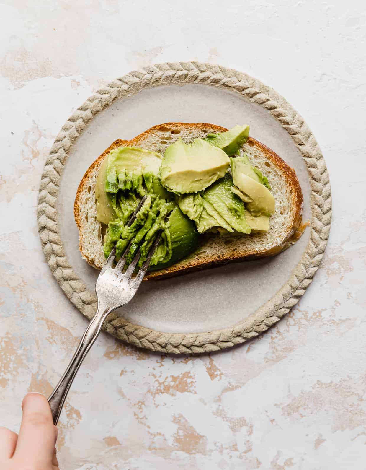 A fork smashing fresh avocado on a slice of toasted bread.