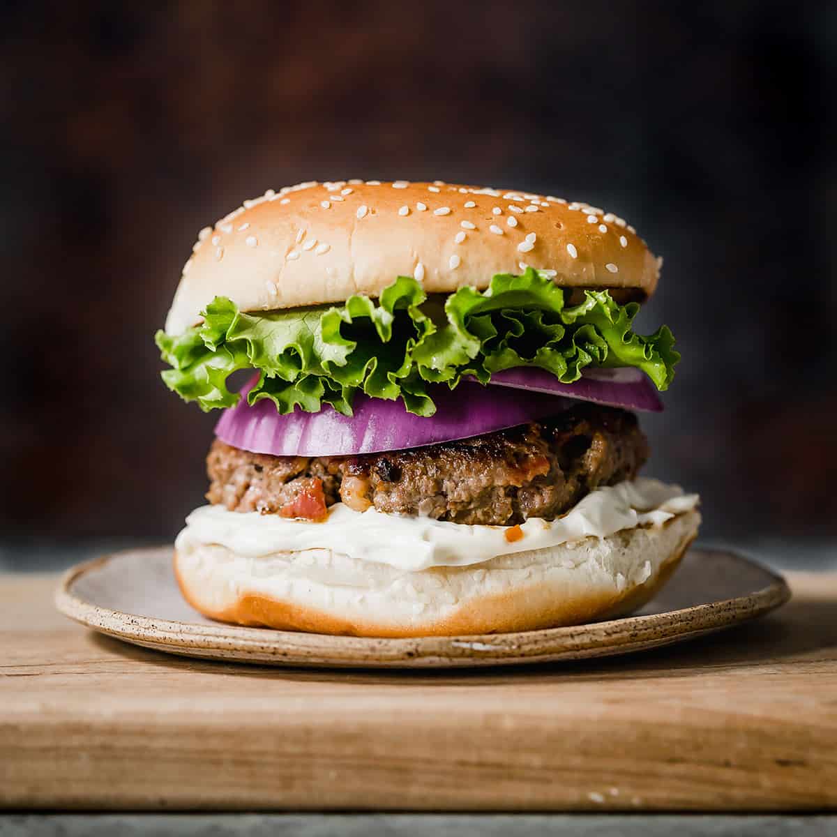 A bacon burger on a cutting board with green lettuce and purple onion on the burger.