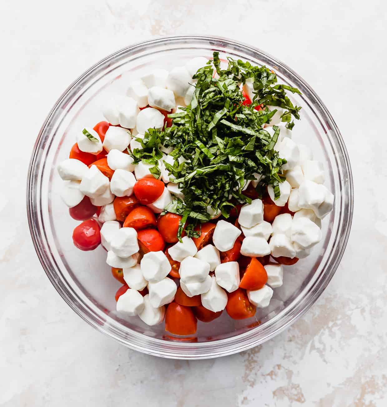 A glass bowl with sliced cherry tomatoes, chopped basil, and mozzarella balls. 