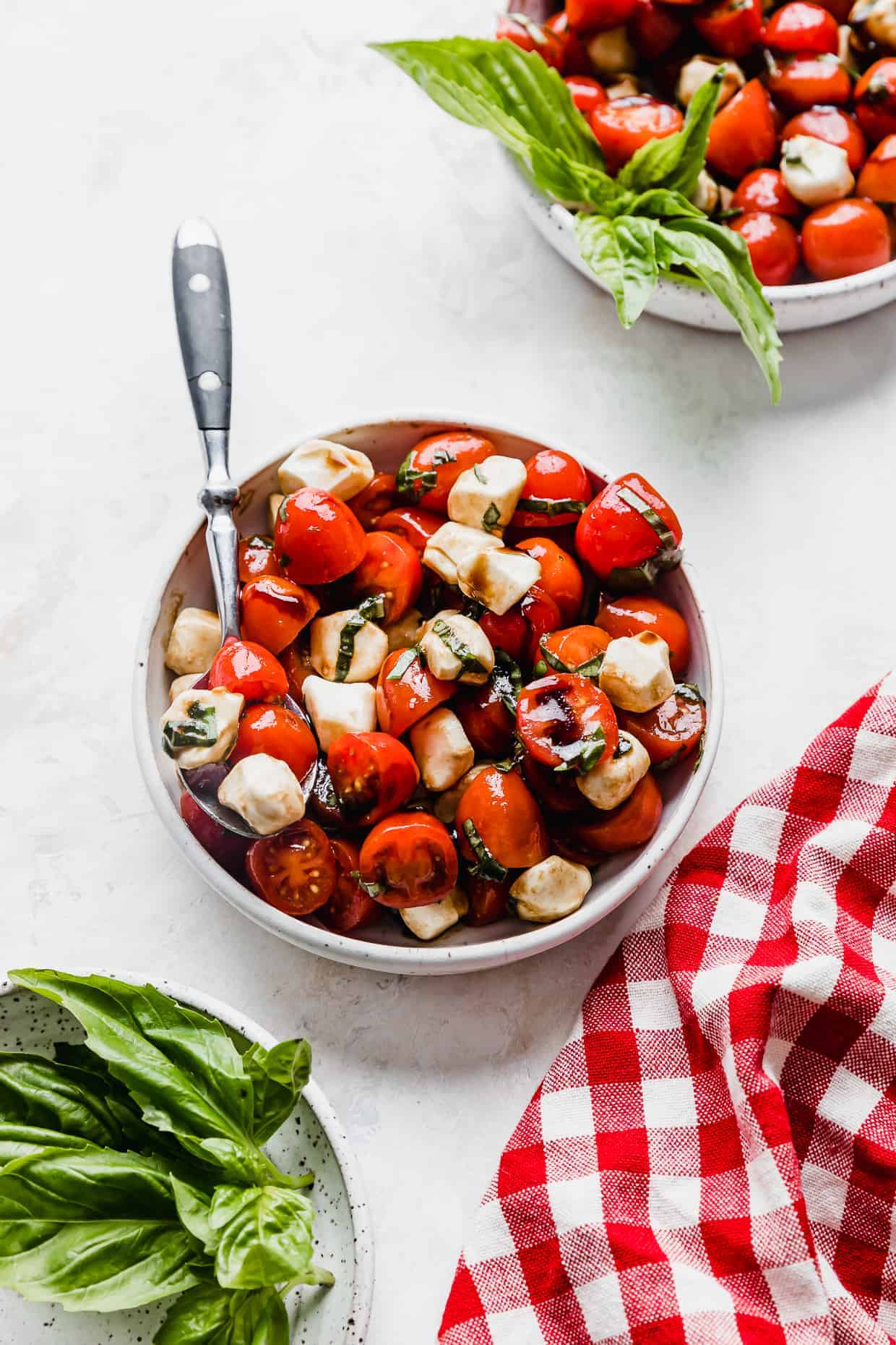 A white bowl with Cherry Tomato Caprese Salad in it, on a white background with fresh basil leaves in the foreground.