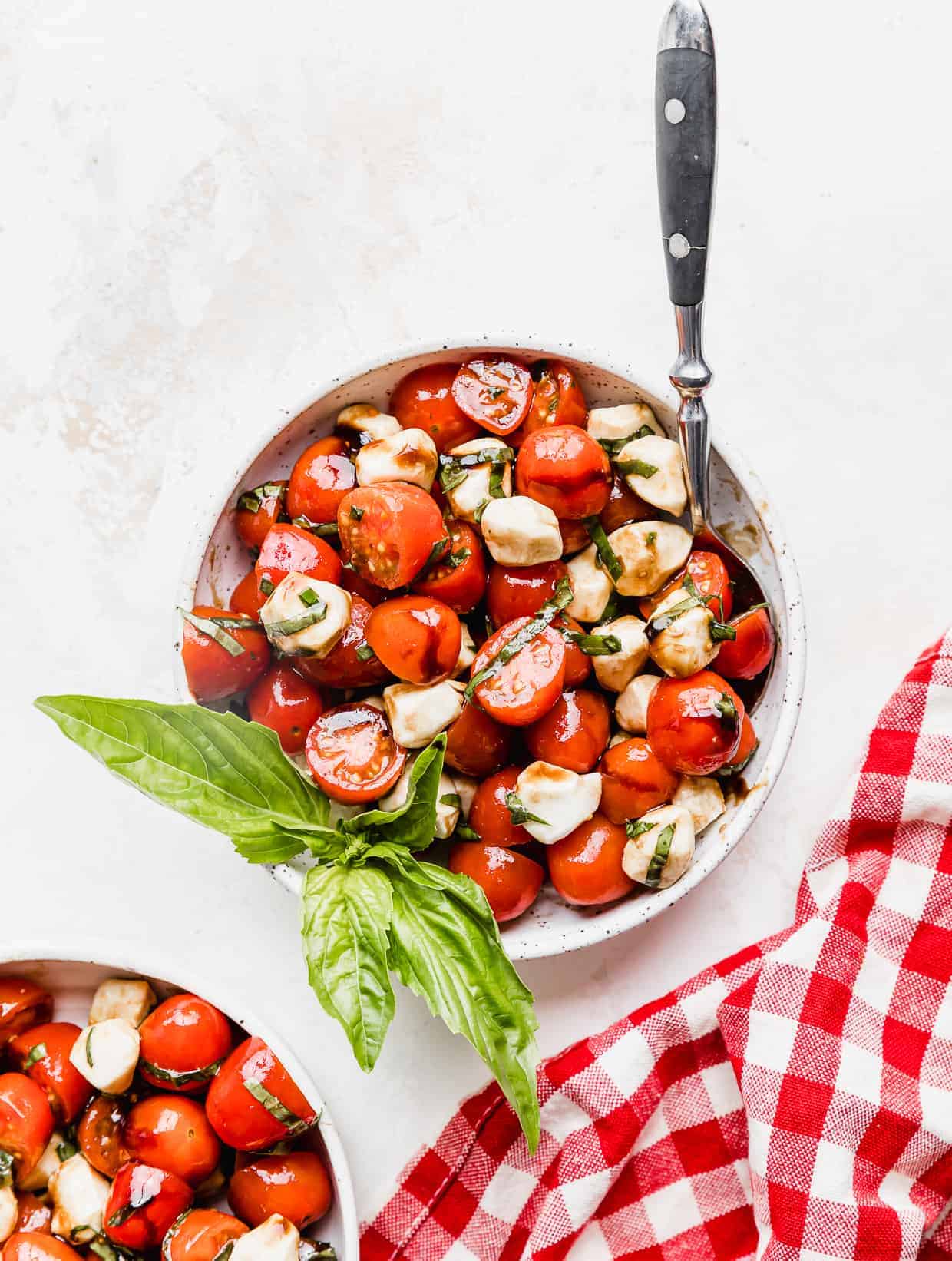 A Cherry Tomato Caprese Salad in a white bowl with a sprig of fresh basil in it with a red checkered napkin next to the bowl. 