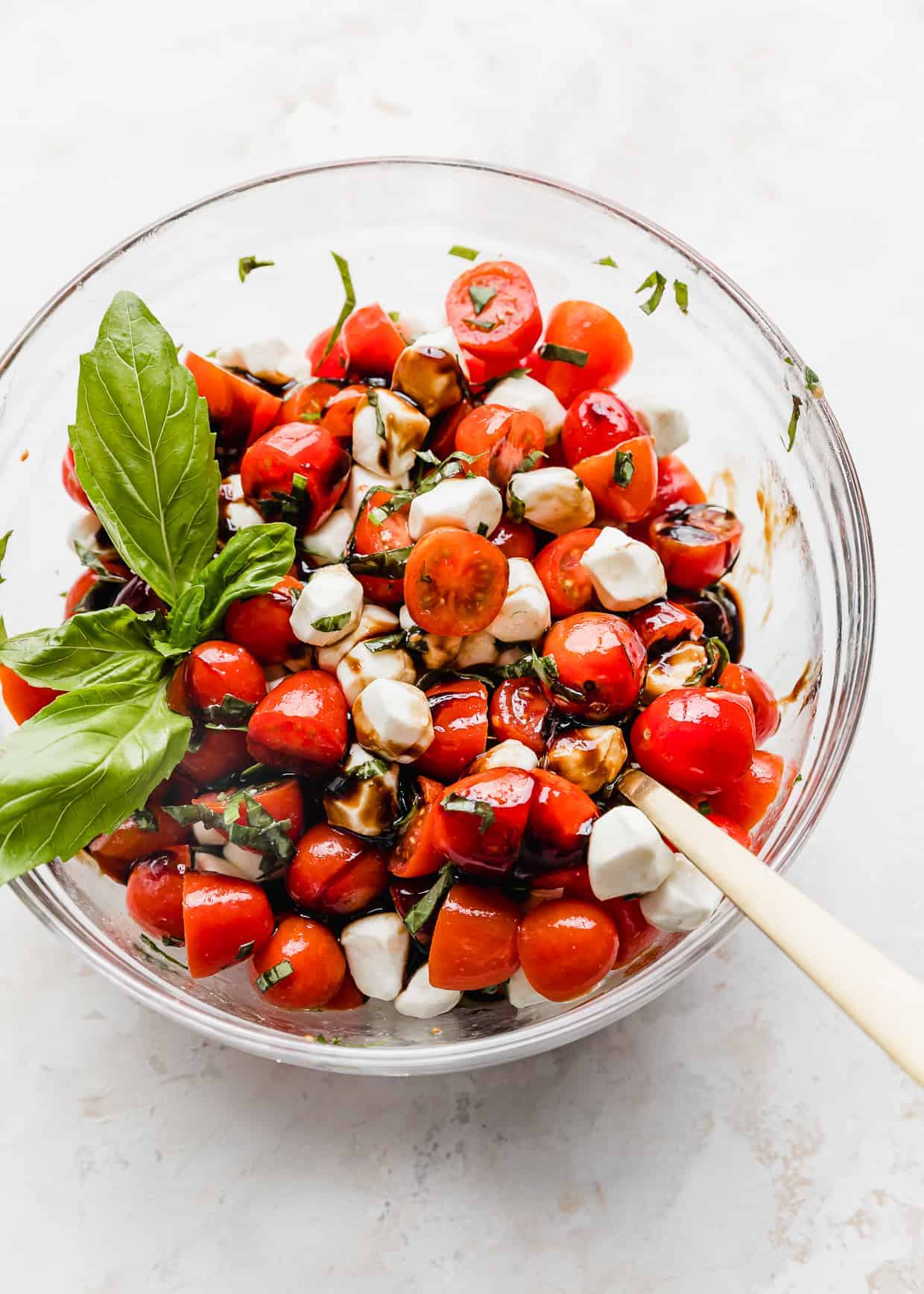 A glass bowl full of red grape or cherry tomatoes with mozzarella balls, chopped basil, and balsamic glaze. 