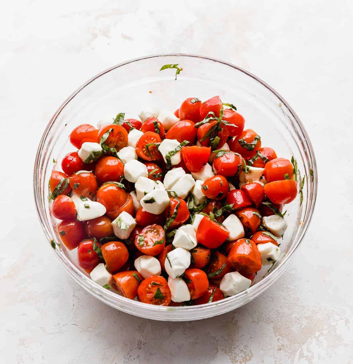 A glass bowl full of red grape tomatoes tossed with mozzarella balls and chopped basil. 