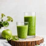 Two glasses full of dark green celery smoothie on a wooden round board.