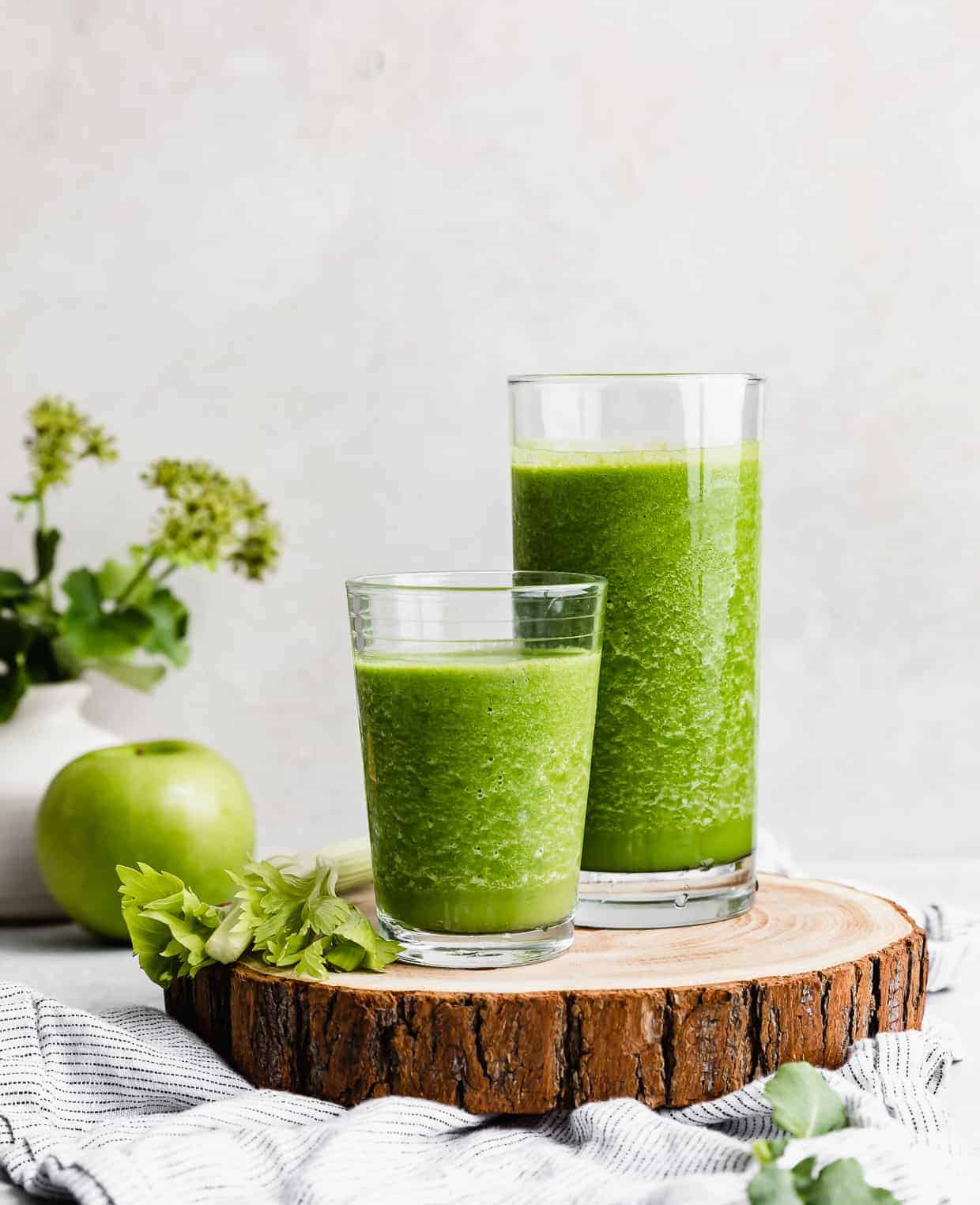 Two glasses full of dark green celery smoothie on a wooden round board.