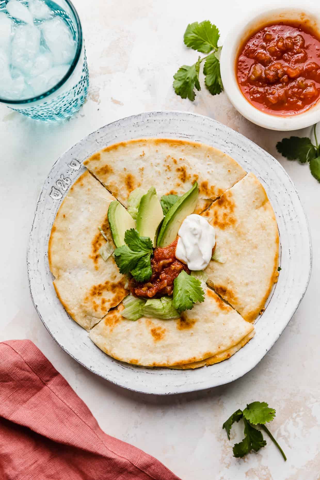 A cheese quesadilla on a white plate that has been cut into 4 wedges, topped with salsa, avocado, sour cream, and lettuce.
