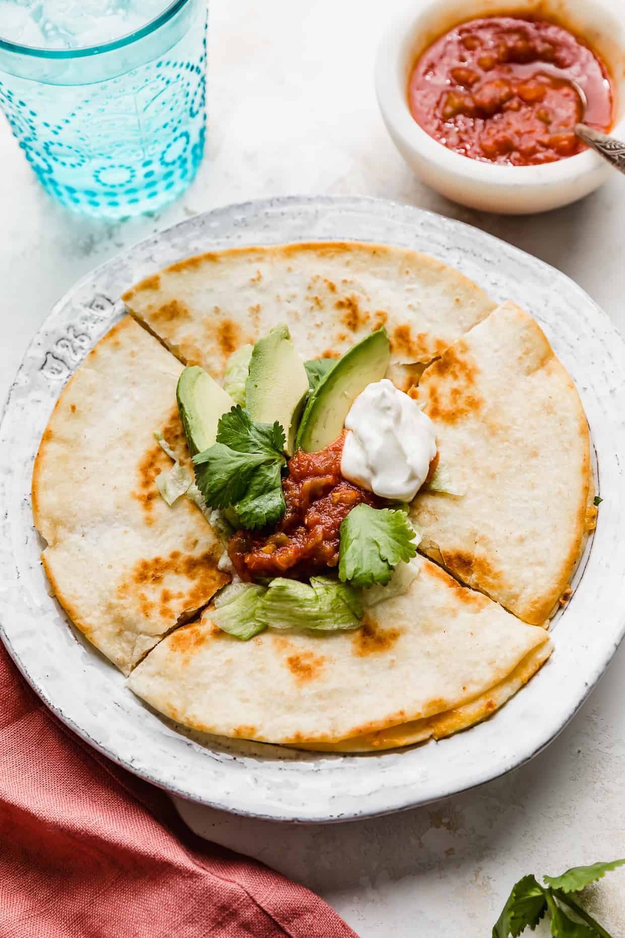 A cheese quesadilla on a white plate topped with avocado, salsa, sour cream, and shredded lettuce.