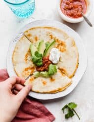 A hand grabbing a slice of a golden brown cheese quesadilla on a white plate that has been topped with avocado, sour cream, and salsa.