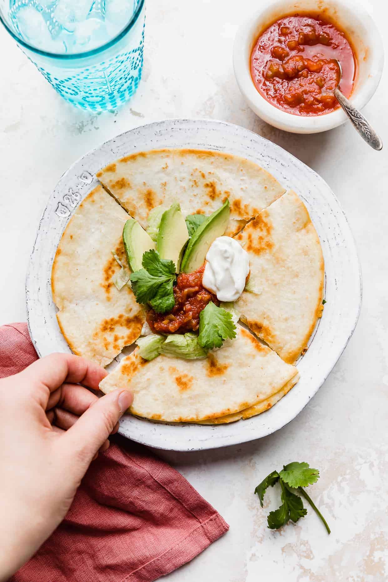 A hand grabbing a slice of a golden brown cheese quesadilla on a white plate that has been topped with avocado, sour cream, and salsa.
