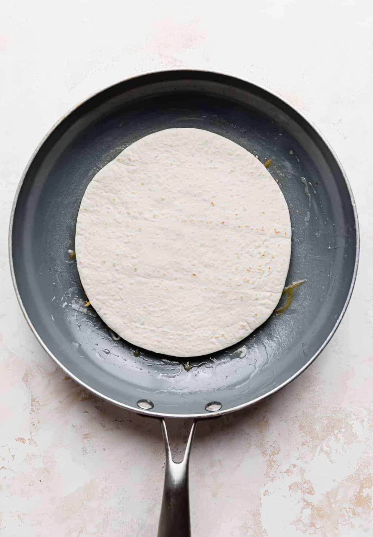 A torilla in a gray frying pan against a white and cream colored background.