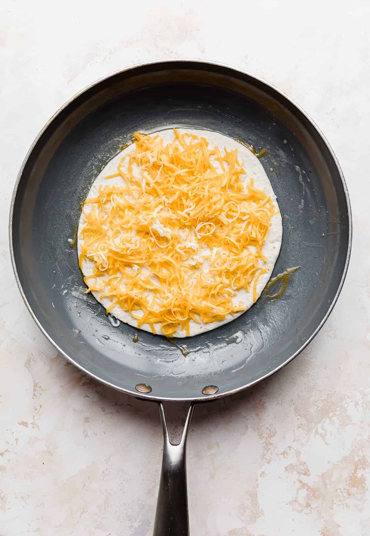 A frying pan with a quesadilla in it that has been topped with shredded cheddar cheese.