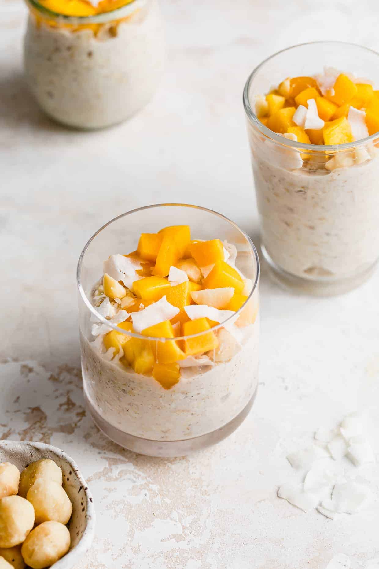 A clear cup full of Mango Overnight Oats topped with diced mangos and shaved coconut flakes.