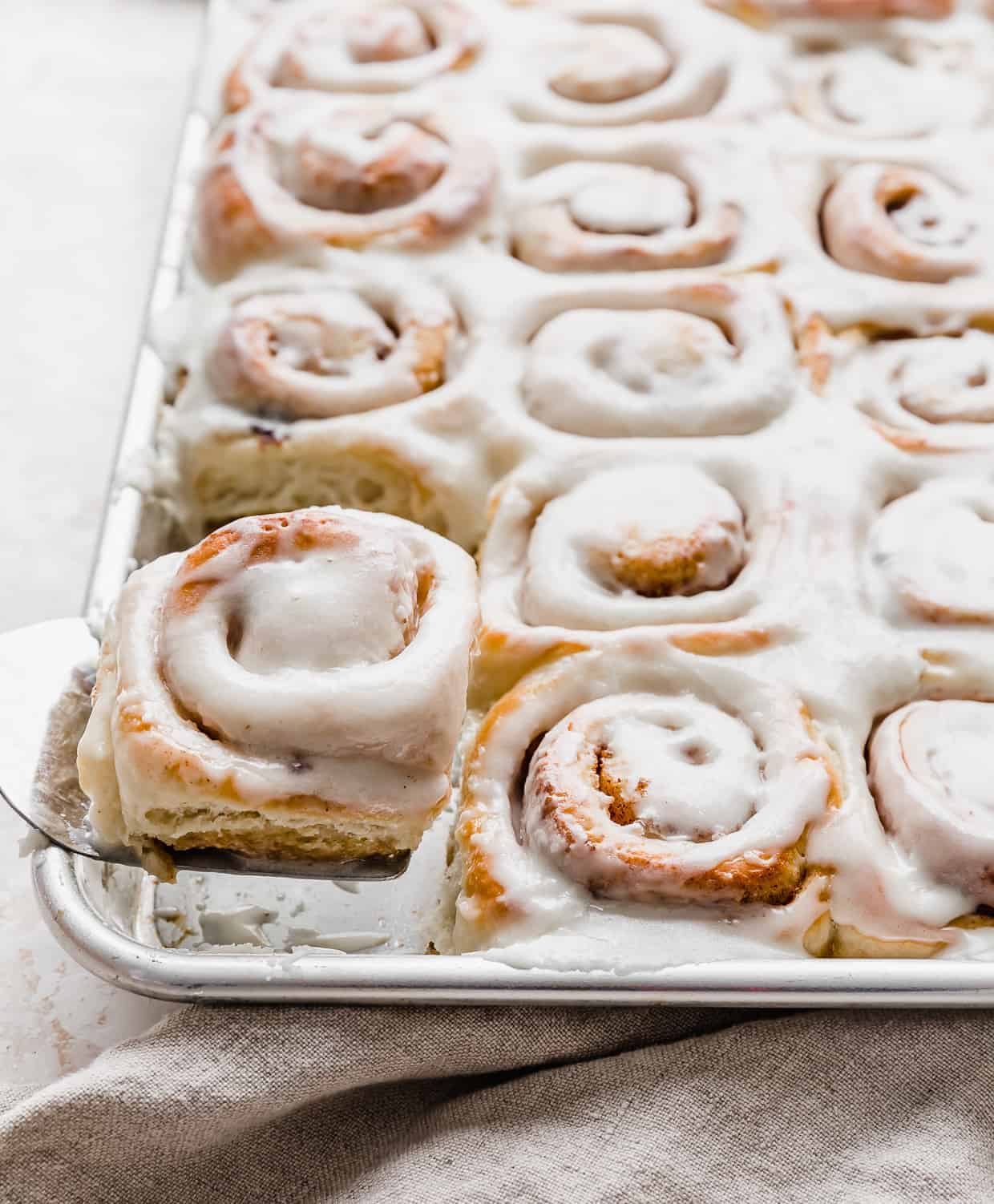 A metal spatula holding up a mini cinnamon roll in front of a baking sheet full of frosted cinnamon rolls. 