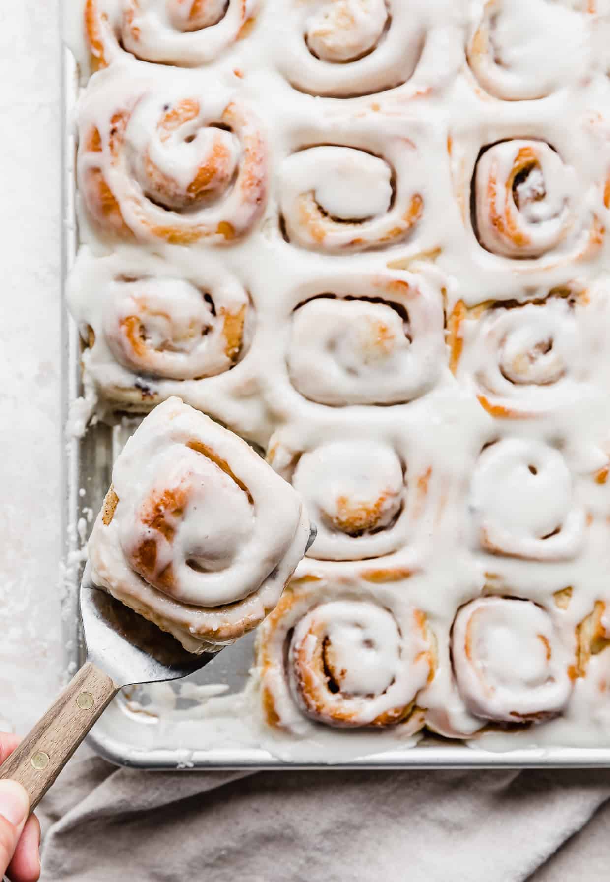 A spatula holding up a mini cinnamon roll with a baking sheet full of others in the background.