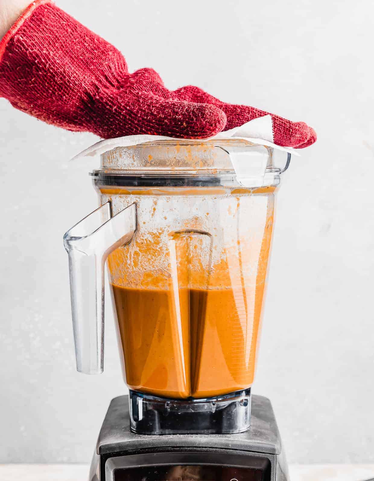 A blender with tomato basil soup in it, with a hot pad covered hand over the blender opening hole.