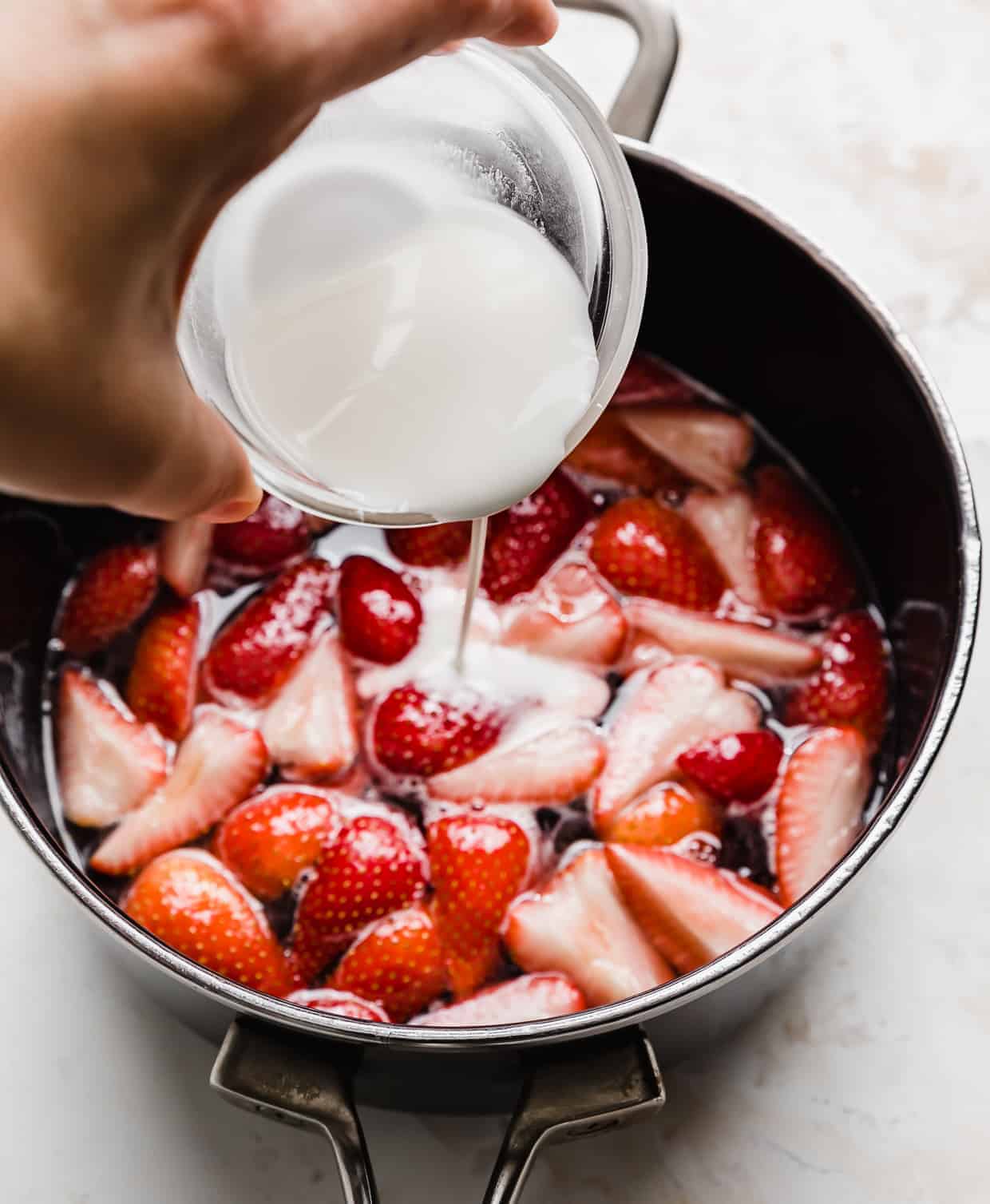 A cornstarch slurry being poured overtop cut strawberries in a pan.