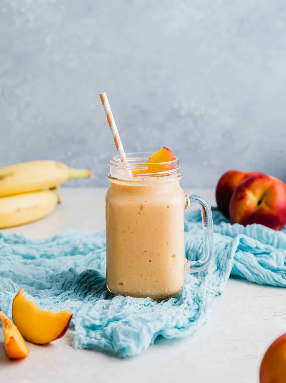 A banana peach smoothie in a glass cup with fresh peaches and bananas in the background.