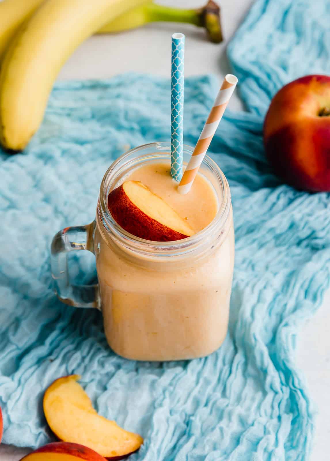 A glass cup filled with a banana peach smoothie topped with a slice of fresh peach.