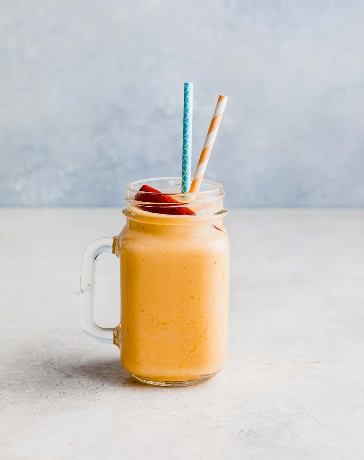 A glass cup filled with an orange colored peaches and banana smoothie against a light blue background.