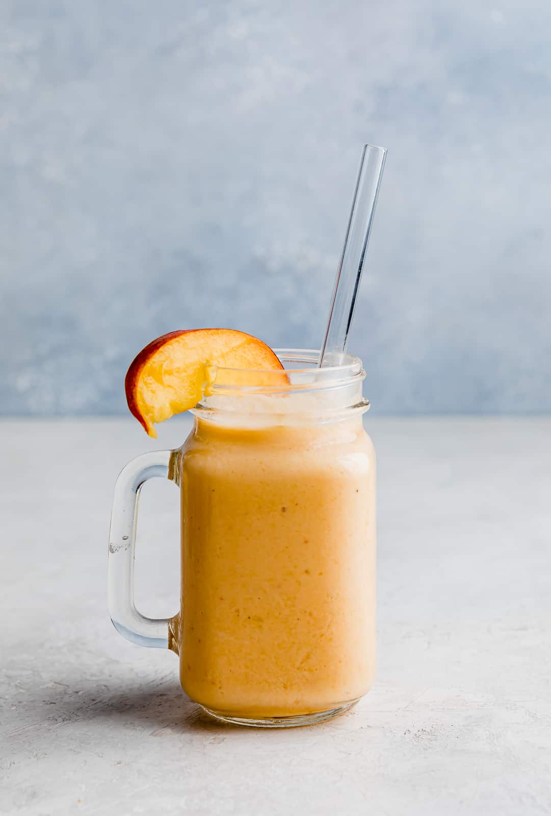 A banana peach smoothie in a glass cup with a fresh peach slice on the rim.