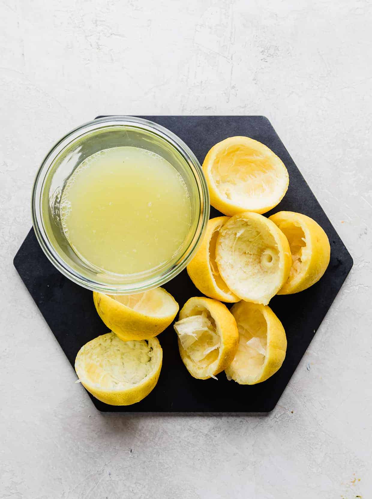 A jar of freshly squeezed lemon juice surrounded by squeezed lemons, on a black hexagon plate.