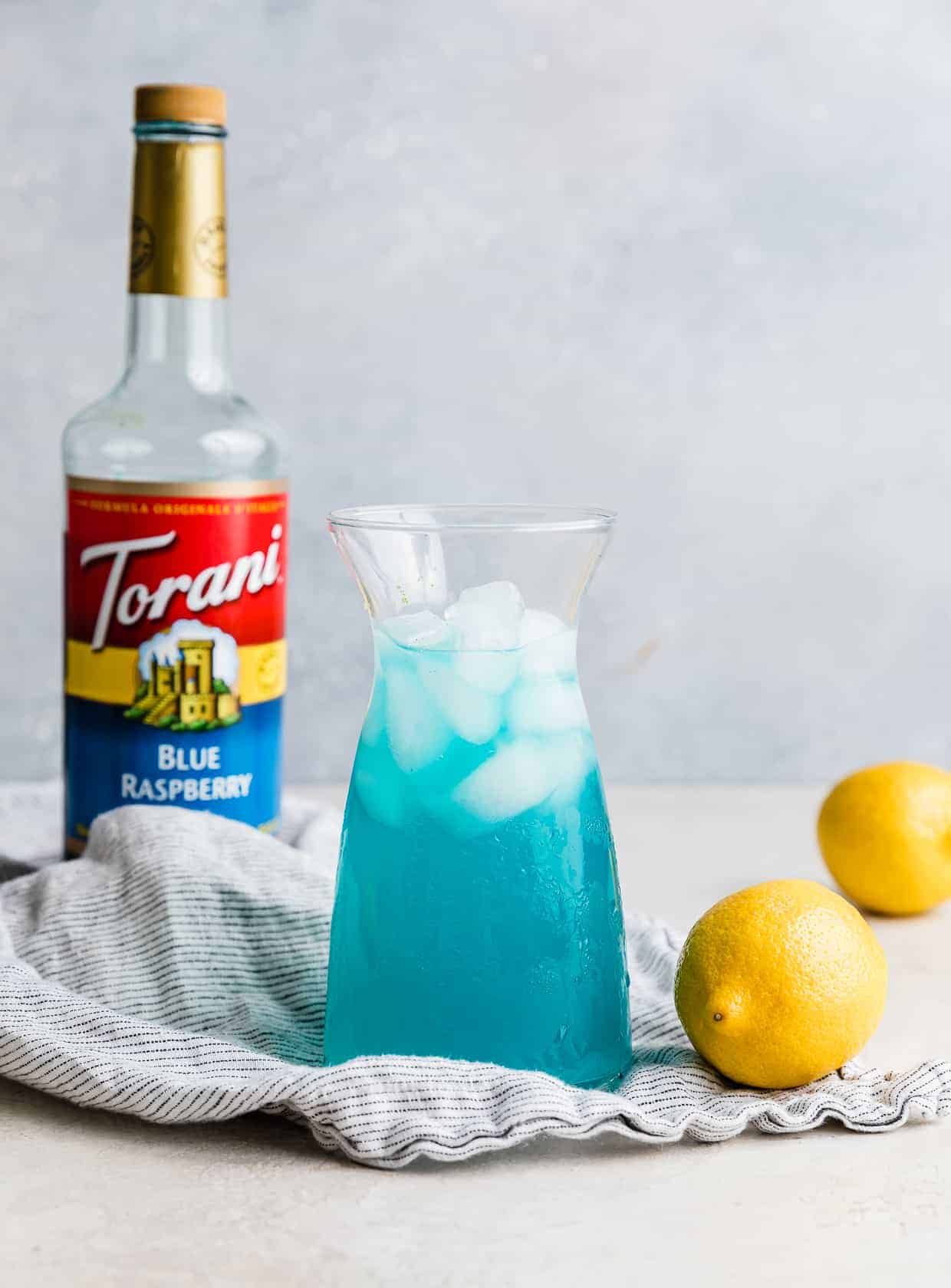 A jar of Blue Raspberry Lemonade against a gray background with a bottle of Torani blue syrup in the background.