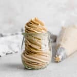 Peanut butter frosting swirly piped into a small glass jar, with piping bag in background.