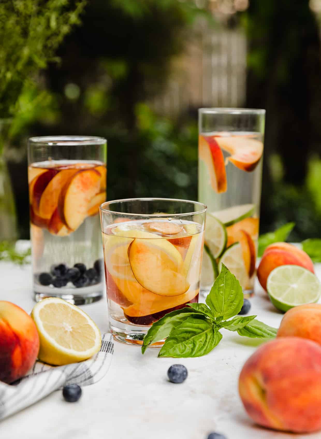 Three glasses of peach infused water with peaches, basil, and lemons surrounding the cups.