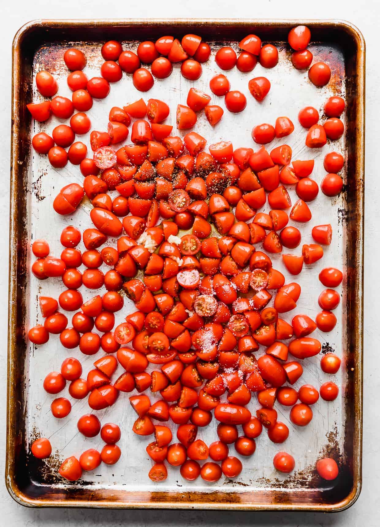 Small red tomatoes on a baking sheet topped with salt, pepper, and fresh garlic.