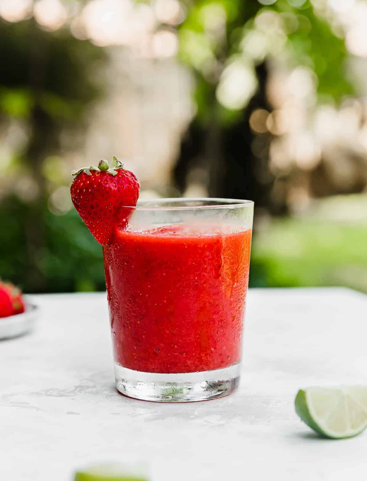 A glass of Virgin Strawberry Daiquiri on a white board with outside greenery in the background.